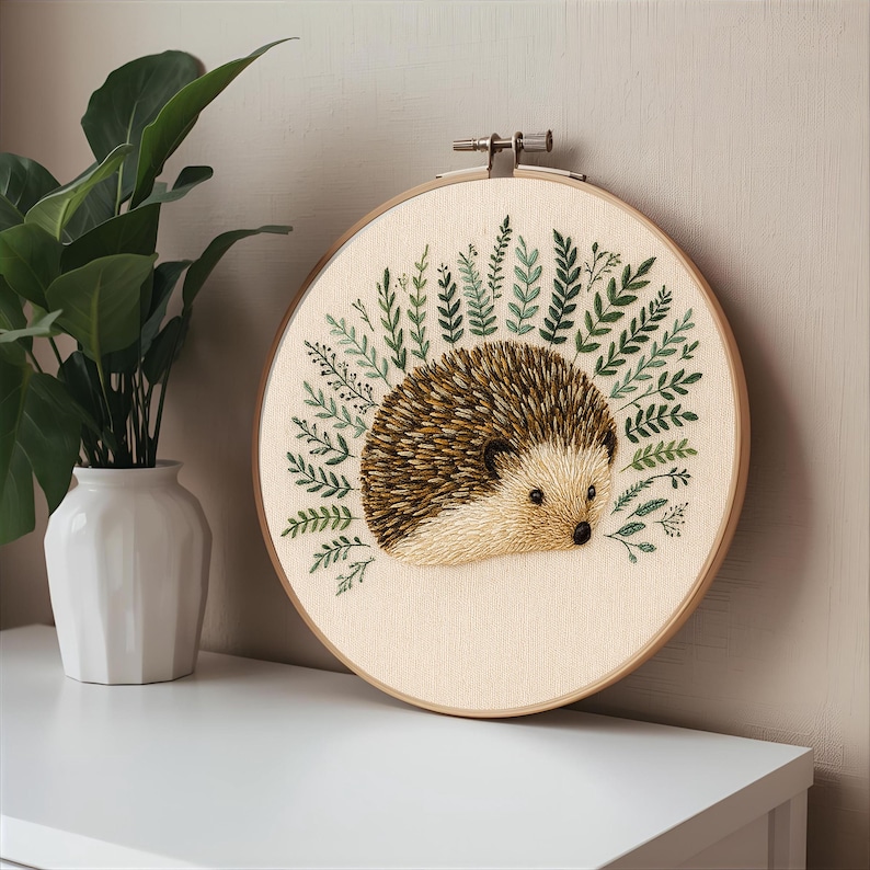 May include: A handmade embroidery hoop featuring a detailed hedgehog design. The hedgehog has brown and tan quills, a light tan face, and is surrounded by green embroidered foliage. The hoop is made of wood and is displayed on a white surface.