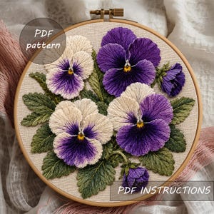May include: A circular wooden embroidery hoop containing a needlepoint design of pansy flowers in shades of purple, cream, and yellow with green leaves on a beige fabric background. The image includes the text PDF pattern and PDF INSTRUCTIONS.