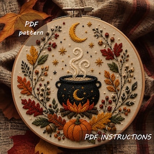 May include: An embroidery hoop featuring a black cauldron over a fire with rising steam, surrounded by autumn leaves, berries, a pumpkin, and a crescent moon. The design includes the text PDF pattern and PDF INSTRUCTIONS on the fabric surface.