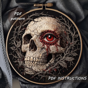 Könnte beinhalten: Gesticktes Schädeldesign in einem schwarzen Stickrahmen. Der Schädel weist Risse und ein rotes Auge auf, umgeben von grauen Zweigen. Der Text "PDF pattern" und "PDF instructions" ist sichtbar.