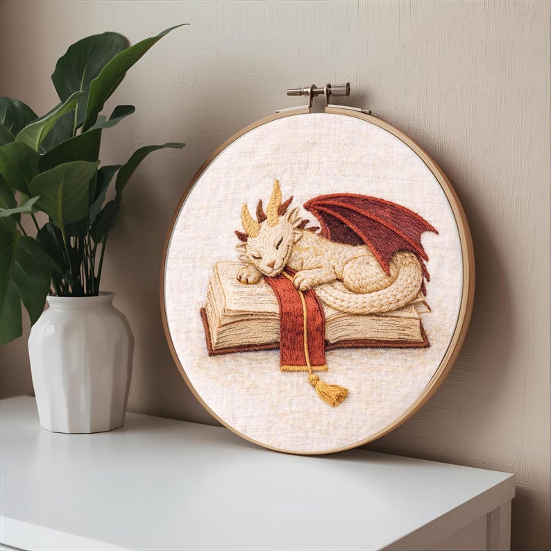 Puede incluir: Obra de arte bordada que representa un drag&oacute;n durmiente sobre una pila de libros. El drag&oacute;n es de color crema con cuernos dorados y alas rojas. Una bufanda roja con una borla dorada descansa sobre los libros. La obra est&aacute; enmarcada en un aro de madera.