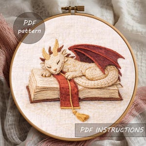 Puede incluir: Dise&ntilde;o de drag&oacute;n bordado en un aro de madera. El drag&oacute;n es de color crema con cuernos marrones y alas rojas, descansando sobre un libro con una bufanda roja. Los textos "PDF pattern" y "PDF instructions" son visibles.