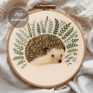 May include: An embroidered hedgehog with brown and tan quills, surrounded by green leaves, is displayed in a wooden embroidery hoop. The text "PDF pattern" is in a circle in the upper left corner. The artwork is on a cream-colored fabric.