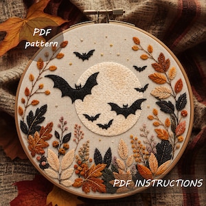 Può includere: Un telaio da ricamo circolare contenente un pannello in tessuto con un disegno ricamato di una grande luna color crema, pipistrelli neri e foglie dai colori autunnali nelle tonalità dell'arancione, marrone e nero. Il testo PDF pattern e PDF INSTRUCTIONS è visibile.
