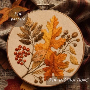 Könnte beinhalten: Ein Stickrahmen mit einem Stoffeinsatz, der ein detailliertes Stickdesign mit herbstlichem Laub zeigt, darunter goldene Eichenblätter, braune farnartige Blätter und kleine rote Beeren. Der Text PDF pattern und PDF INSTRUCTIONS ist zu sehen.