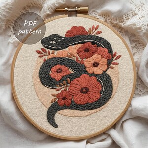 Op de afbeelding: Geborduurde hoepelkunst met een grijze slang die zich rond koraal- en perzikroze bloemen en bladeren wikkelt. Het ontwerp is geplaatst op een beige stof in een houten borduurring. Tekst in de linkerbovenhoek luidt "PDF pattern".