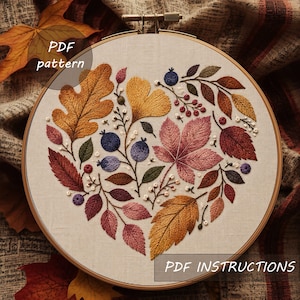 May include: An embroidery hoop containing a fabric panel with a heart-shaped arrangement of stitched autumn leaves and berries in shades of gold, rust, pink, and blue. The text "PDF pattern" and "PDF INSTRUCTIONS" are visible on the image.