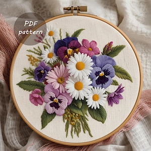 May include: Embroidered floral design featuring daisies, pansies, and other flowers in shades of white, pink, purple, and yellow. The embroidery is set in a wooden hoop, with the text "PDF pattern" in a circle.