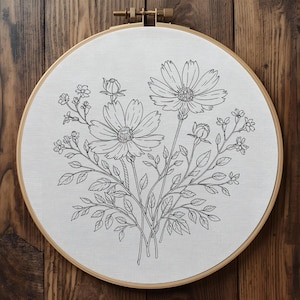 Puede incluir: Un bastidor de bordado floral con un detallado dibujo lineal en blanco y negro de flores de cosmos, capullos y follaje. El dise&ntilde;o est&aacute; centrado en una tela blanca, dentro de un bastidor de madera redondo con cierre de lat&oacute;n.
