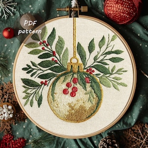 Könnte beinhalten: Gesticktes Weihnachtsornament-Design in einem Holzreifen. Das Ornament ist goldfarben mit grünen Blättern und roten Beeren. Der Text "PDF pattern" befindet sich in einem Kreis oben links. Der Hintergrund ist ein dunkelgrüner Stoff.