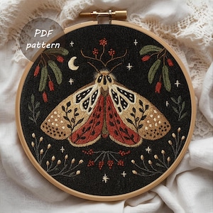Puede incluir: Diseño de polilla bordada sobre tela negra, en un aro de madera. La polilla presenta detalles en dorado, rojo y marrón, con motivos florales y estrellas. El texto "PDF pattern" es visible.