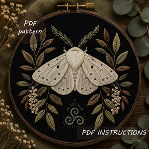 Può includere: Un telaio da ricamo con sfondo in tessuto nero e una falena color crema al centro, circondata da foglie ricamate verdi e marroni e piccoli fiori bianchi. Sotto la falena c'è un simbolo a spirale. Testo: PDF pattern e PDF INSTRUCTIONS.