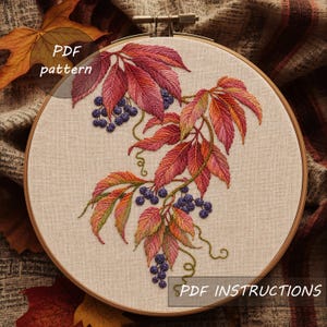May include: An embroidery hoop containing a beige fabric with a hand-stitched design of autumn leaves in shades of red, orange, and yellow, paired with clusters of dark purple berries and green tendrils. The text PDF pattern and PDF INSTRUCTIONS appear.