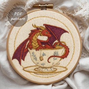 May include: Embroidered hoop art featuring a red and gold dragon nestled in a teacup with blue floral details. The dragon has purple wings and is set against a beige background. The text "PDF pattern" is in a circle at the top.
