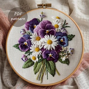 May include: Embroidered floral hoop art featuring a bouquet of purple pansies, white daisies, and green leaves. The embroidery is set in a wooden hoop with the text "PDF pattern" in a circle.