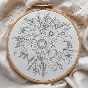 May include: A floral embroidery hoop featuring a detailed black and white design of sunflowers, leaves, and berries. The embroidery is set against a white fabric, framed by a natural wood hoop. The design is intricate and delicate.