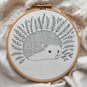May include: A hand-stitched embroidery hoop featuring a detailed black and white hedgehog design. The hedgehog is surrounded by leafy sprigs, all set against a white fabric background. The wooden hoop adds a natural touch to this charming artwork.