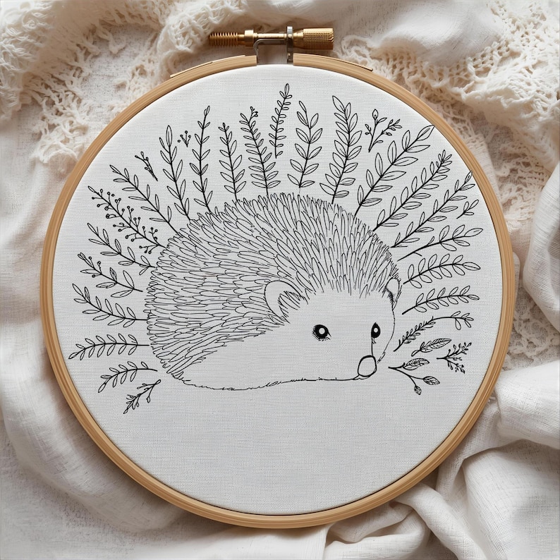 May include: A hand-stitched embroidery hoop featuring a detailed black and white hedgehog design. The hedgehog is surrounded by leafy sprigs, all set against a white fabric background. The wooden hoop adds a natural touch to this charming artwork.