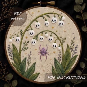 Op de afbeelding: Een borduurring met beige stof waarop lelietjes-van-dalen in de vorm van witte schedels, een paarse spin, spinnenwebben en kleine gouden sterren zijn geborduurd. De tekst PDF pattern en PDF INSTRUCTIONS is zichtbaar.