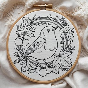 May include: A black and white embroidery hoop featuring a detailed illustration of a bird perched amidst leaves and berries. The design is set against a white fabric background, framed by a natural wood hoop.