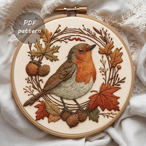 May include: Embroidered artwork of a robin bird surrounded by autumn leaves and acorns, set within a wooden embroidery hoop. The bird has a red breast, brown wings, and a gray belly. The text "PDF pattern" is visible.