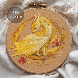 May include: Embroidered dragon design in shades of yellow and gold, set within a wooden embroidery hoop. The dragon features detailed stitching, with red eyes and a wing. The design includes autumn leaves and the text "PDF pattern".