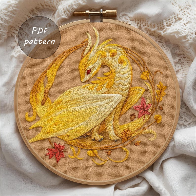 May include: Embroidered dragon design in shades of yellow and gold, set within a wooden embroidery hoop. The dragon features detailed stitching, with red eyes and a wing. The design includes autumn leaves and the text "PDF pattern".