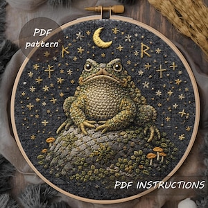 May include: A circular embroidery hoop with a detailed frog design. The frog is green and brown, sitting on a mossy rock, under a crescent moon and stars. The background is dark blue. Text reads "PDF pattern" and "PDF instructions".