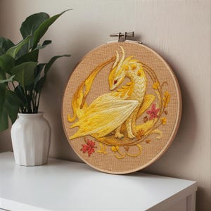 May include: Embroidered artwork of a yellow dragon with red eyes, set against a tan background within a wooden embroidery hoop. The dragon is surrounded by autumn leaves and vines, creating a fantasy theme. The artwork is displayed on a white surface.