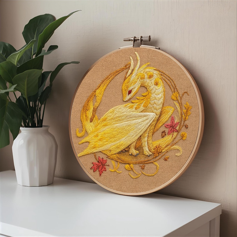 May include: Embroidered artwork of a yellow dragon with red eyes, set against a tan background within a wooden embroidery hoop. The dragon is surrounded by autumn leaves and vines, creating a fantasy theme. The artwork is displayed on a white surface.