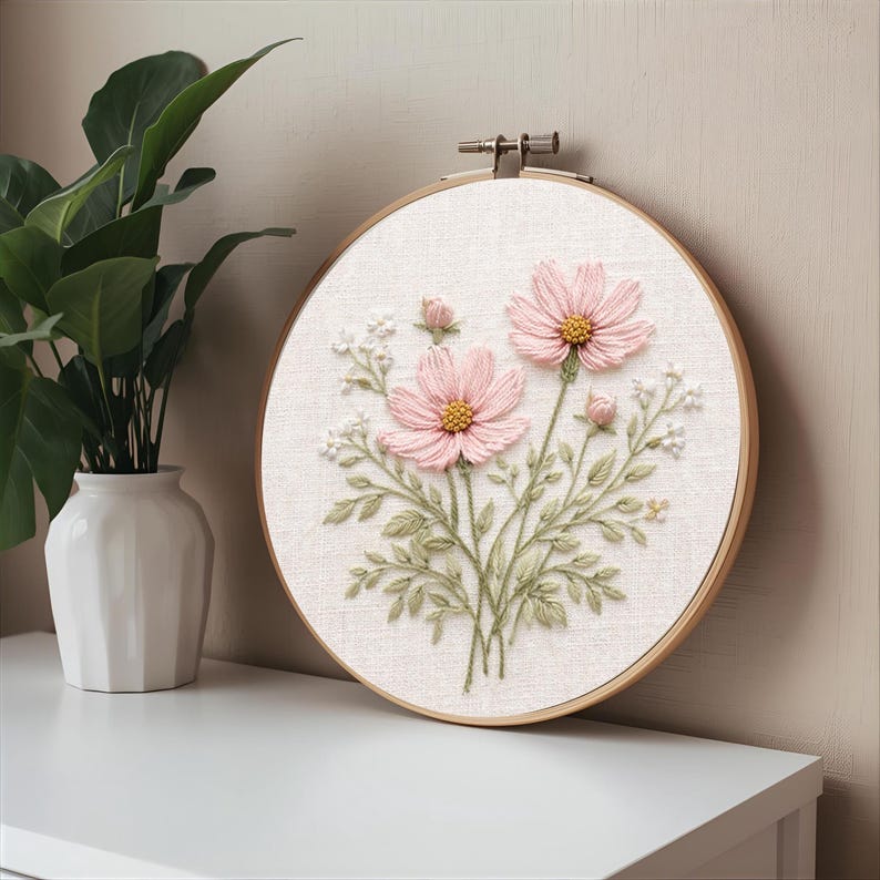 Puede incluir: Un bastidor de bordado redondo con un dise&ntilde;o floral. Dos flores de cosmos rosas con centros amarillos son el punto focal, rodeadas de hojas verdes y peque&ntilde;as flores blancas. El bordado est&aacute; sobre una tela color crema, enmarcado por un aro de madera.