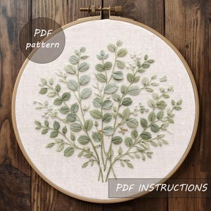 May include: An embroidery hoop featuring a detailed floral design with green leaves and small white flowers. The design is centered on a cream-colored fabric, set within a wooden hoop. Text on the image reads "PDF pattern" and "PDF instructions".
