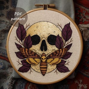 May include: Embroidered skull and moth design in a wooden hoop. The skull is beige with black eye sockets and nose. The moth has yellow and brown wings. Purple leaves surround the skull. The text "PDF pattern" is in the upper left corner.