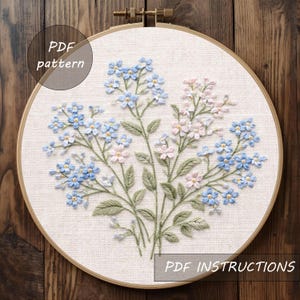 Könnte beinhalten: Ein Stickrahmen mit einem floralen Design, das blaue und rosa Blumen und grüne Blätter zeigt. Die Texte "PDF pattern" und "PDF instructions" sind sichtbar. Die Stickerei ist in einem Holzrahmen befestigt.