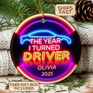 May include: A round Christmas ornament with a car silhouette and the text "THE YEAR I TURNED DRIVER OLIVIA 2025" in neon-style lettering. The ornament has a gold string for hanging and is surrounded by festive decorations.