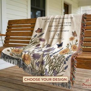 Puede incluir: Una manta beige con un diseño floral y mariposas, sobre un columpio de madera. La manta tiene el texto "NEVER UNDERESTIMATE THE difference...HAPPY RETIREMENT Samantha". La manta tiene un borde con flecos y está decorada con flores amarillas, moradas y azules.