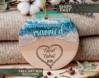 Puede incluir: Un adorno de cerámica redondo con una escena de playa con olas azules rompiendo en una orilla arenosa. El texto dice Our First Christmas married, con un contorno de corazón que contiene Name1 & Name2 2026. Incluye un cordón dorado.