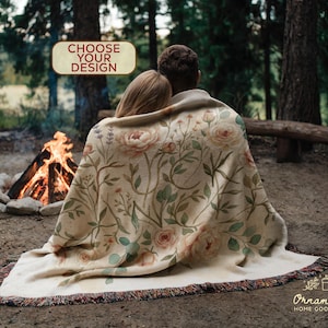 May include: A beige floral throw blanket with a fringed edge, draped over two people sitting near a campfire. The blanket features a pattern of pink roses and green leaves. The text "CHOOSE YOUR DESIGN" is in a tan box.