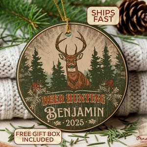 May include: Round wooden Christmas ornament with a deer illustration, pine trees, and the text "Deer Hunting Benjamin 2025". The ornament has a rustic, vintage style with a gold hanging string.