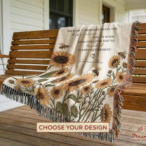 May include: A beige throw blanket with a sunflower and bee design, draped over a wooden swing. The blanket has a fringed edge and text that reads "NEVER UNDERESTIMATE THE difference YOU'VE MADE, AND THE LIVES YOU'VE touched HAPPY RETIREMENT Samantha."