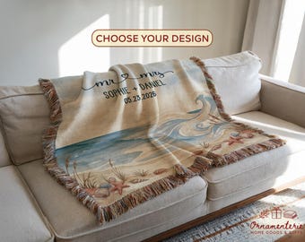 Può includere: Una coperta beige tessuta con un design a scena di spiaggia, con onde oceaniche, sabbia e stelle marine. La coperta ha un bordo con frange e include il testo "mr & mrs SOPHIE + DANIEL 05.23.2026".