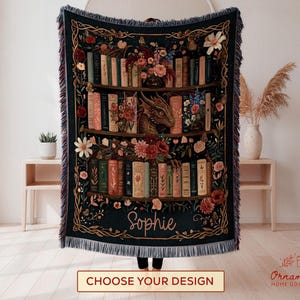 May include: A dark teal woven throw blanket with a bookshelf design, featuring books, flowers, and a dragon. The name "Sophie" is written in cursive. The blanket has a fringed edge and the text "CHOOSE YOUR DESIGN" is at the bottom.