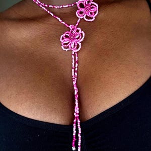 Peut inclure: Un collier style lariat composé de petites perles de rocaille dans des tons rose, magenta et blanc. Le design présente deux motifs en forme de fleurs réalisés en perles, avec de longs brins perlés pendants descendant de la fleur centrale.