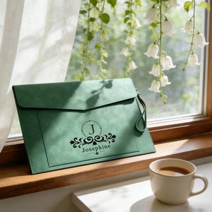 Puede incluir: Un bolso de mano verde salvia con solapa y correa de muñeca. Presenta un monograma negro con la letra "J" y el nombre "Josephine". Una taza de café está cerca.