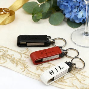 Può includere: Tre chiavette USB in pelle nera, rossa e bianca, ciascuna con un portachiavi argentato. L'unità nera ha il nome "Noah" in corsivo, la rossa "Kyman" e la bianca "H|L".