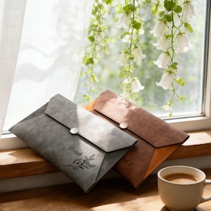 Puede incluir: Dos fundas de documentos de cuero estilo sobre en gris y marrón, con cierre de botón. La funda gris tiene el nombre "Dinah" y un diseño floral. Una taza de café está cerca.