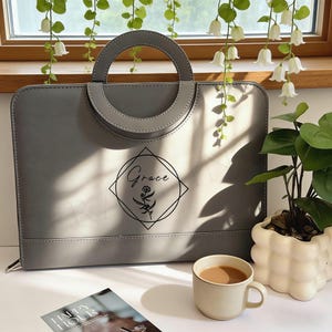 Puede incluir: Una bolsa gris para portátil con un asa circular y el nombre "Grace" en un diseño en forma de diamante. También se ve una taza pequeña de café y una planta en maceta.