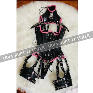 May include: Black leather corset with pink trim and accents. The corset features multiple straps, buckles, and metal hardware. The text "IRON ROOT LEATHER" is visible across the image.