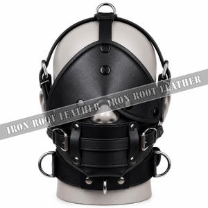 Può includere: Un'imbracatura e maschera per la testa in pelle nera con una copertura solida per gli occhi, un dispositivo di restrizione della bocca con fibbie e molteplici anelli a D e borchie in metallo argentato. Il testo IRON ROOT LEATHER è stampato.