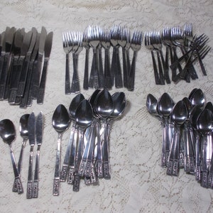 May include: A set of silverware in a silver finish. The set includes forks, knives, and spoons. The silverware is arranged in rows on a white tablecloth.
